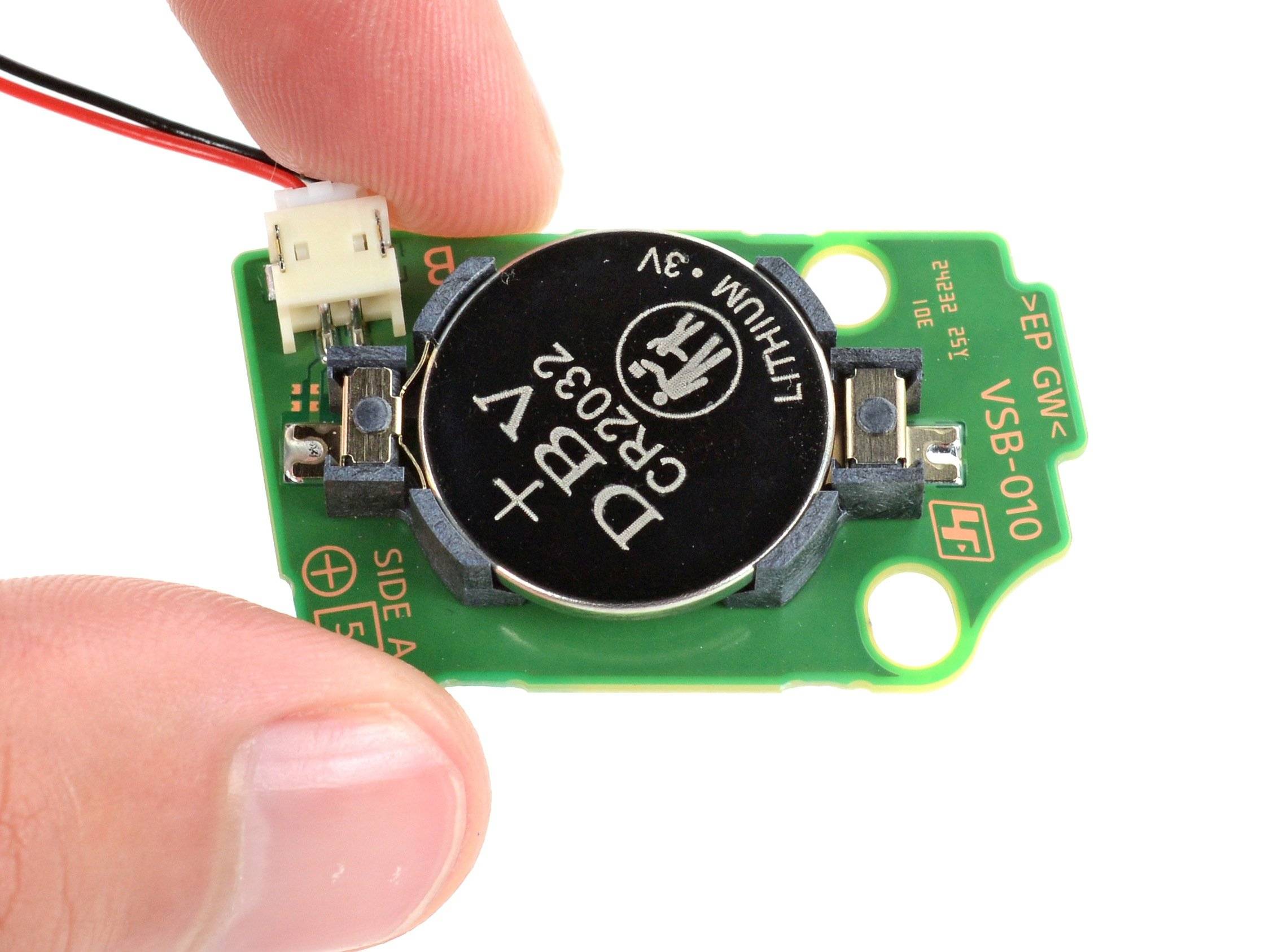 Close up of a PlayStation 5 CMOS board showing the removable CR2032 coin battery held in fingers